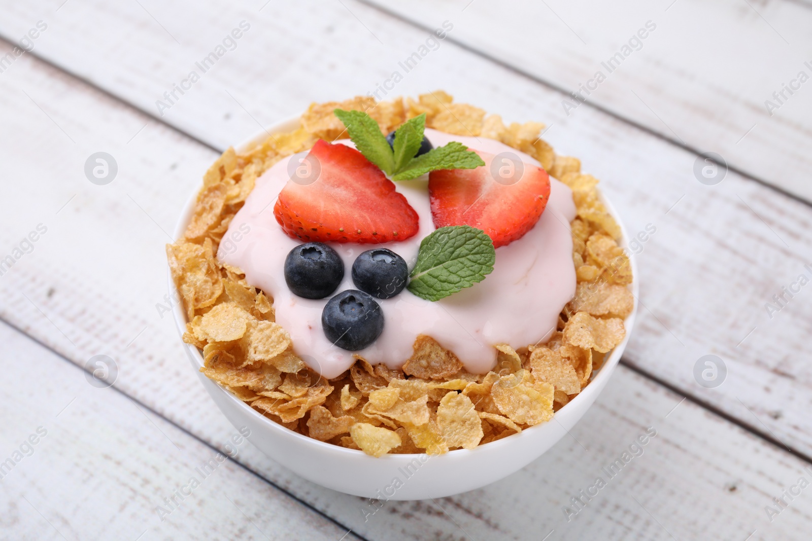 Photo of Delicious crispy cornflakes, yogurt and fresh berries in bowl on white wooden table, closeup. Healthy breakfast
