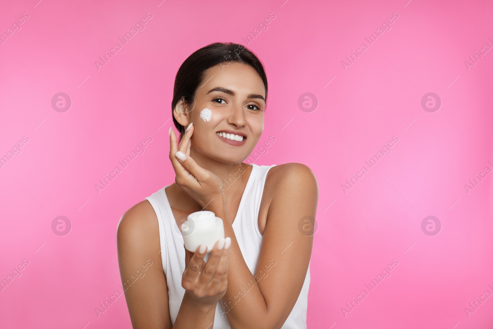 Photo of Young woman applying facial cream on pink background