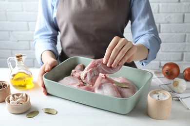 Photo of Woman putting raw rabbit meat into baking dish at white wooden table, closeup