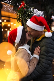 Happy couple in Santa hats kissing under mistletoe bunch outdoors, bokeh effect