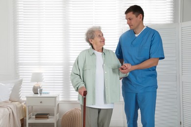 Photo of Caregiver assisting senior woman with walking cane indoors. Home health care service