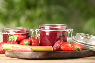 Photo of Tasty rhubarb jam in jars, stems and strawberries on wooden table against blurred background