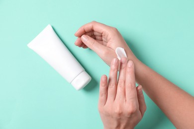 Photo of Woman with tube applying cosmetic cream onto her hand on turquoise background, top view