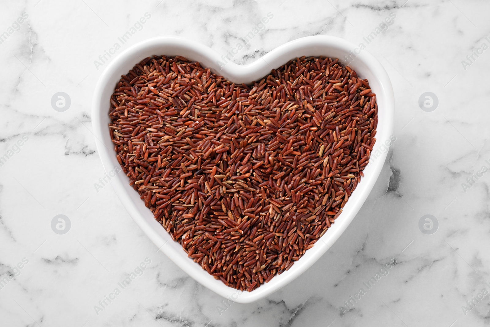 Photo of Brown rice in heart shaped bowl on white marble table, top view