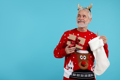 Photo of Senior man in Christmas sweater, reindeer headband holding gift and stocking on light blue background. Space for text