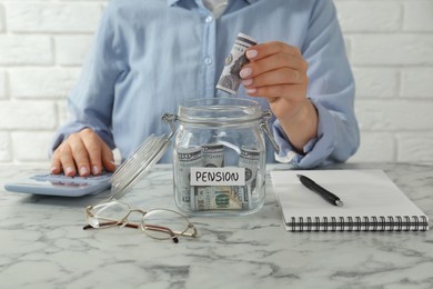 Photo of Pension savings. Woman putting dollar banknote into glass jar with money at marble table, closeup