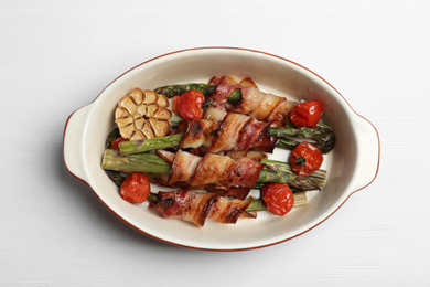 Photo of Oven baked asparagus wrapped with bacon in ceramic dish on white wooden table, top view