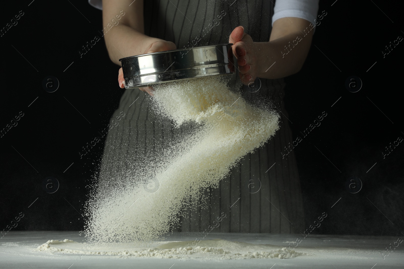 Photo of Woman sieving flour at table against black background, closeup