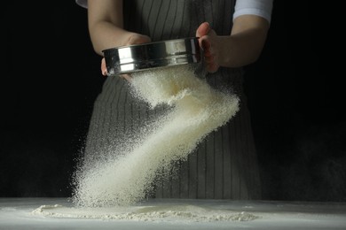 Photo of Woman sieving flour at table against black background, closeup