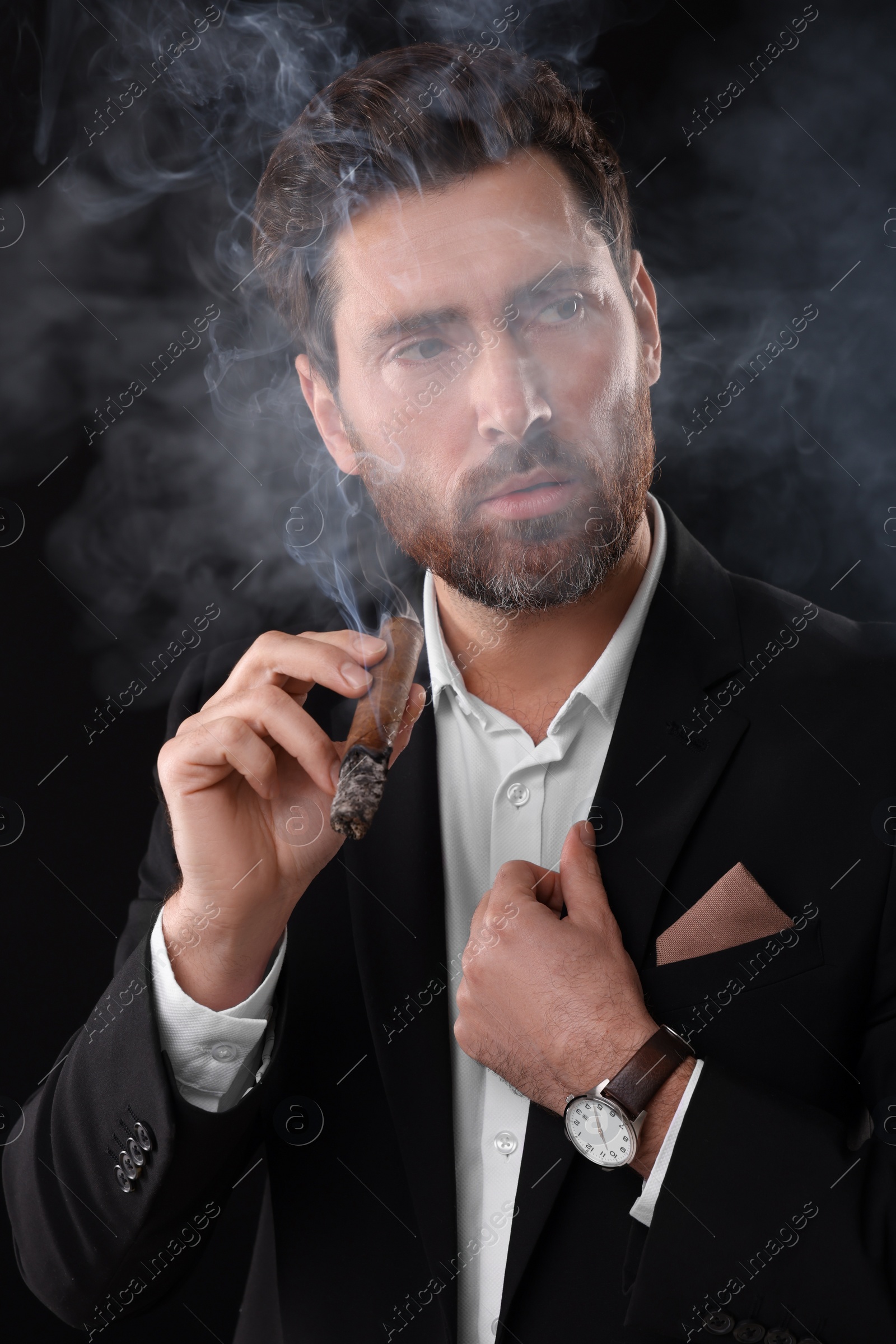 Photo of Handsome man in elegant suit smoking cigar on black background