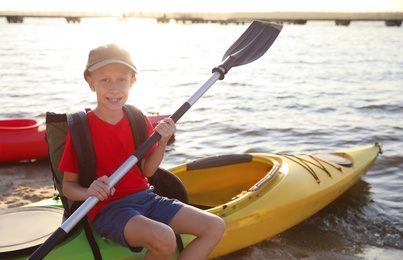 Photo of Happy boy with paddle sitting on kayak near river. Summer camp activity
