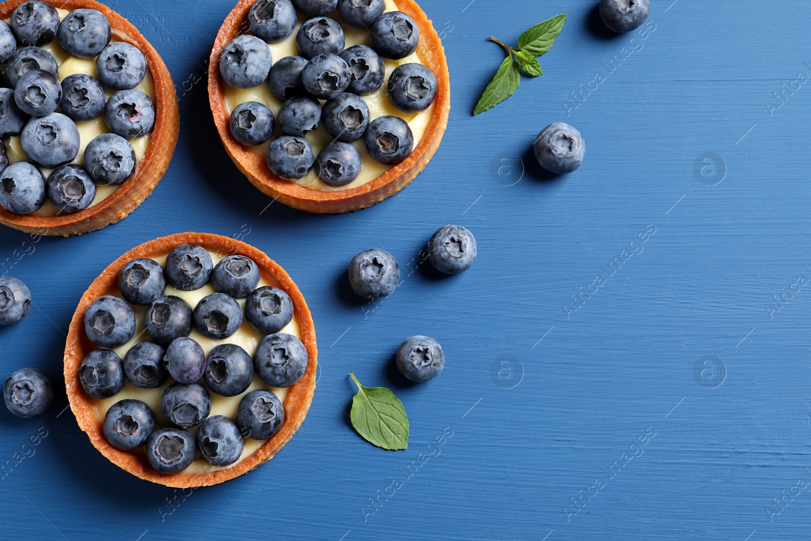 Photo of Tartlets with fresh blueberries and mint on blue wooden table, flat lay with space for text. Delicious dessert
