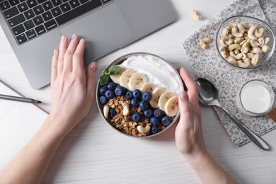 Woman with bowl of tasty granola working with laptop at white wooden table, top view