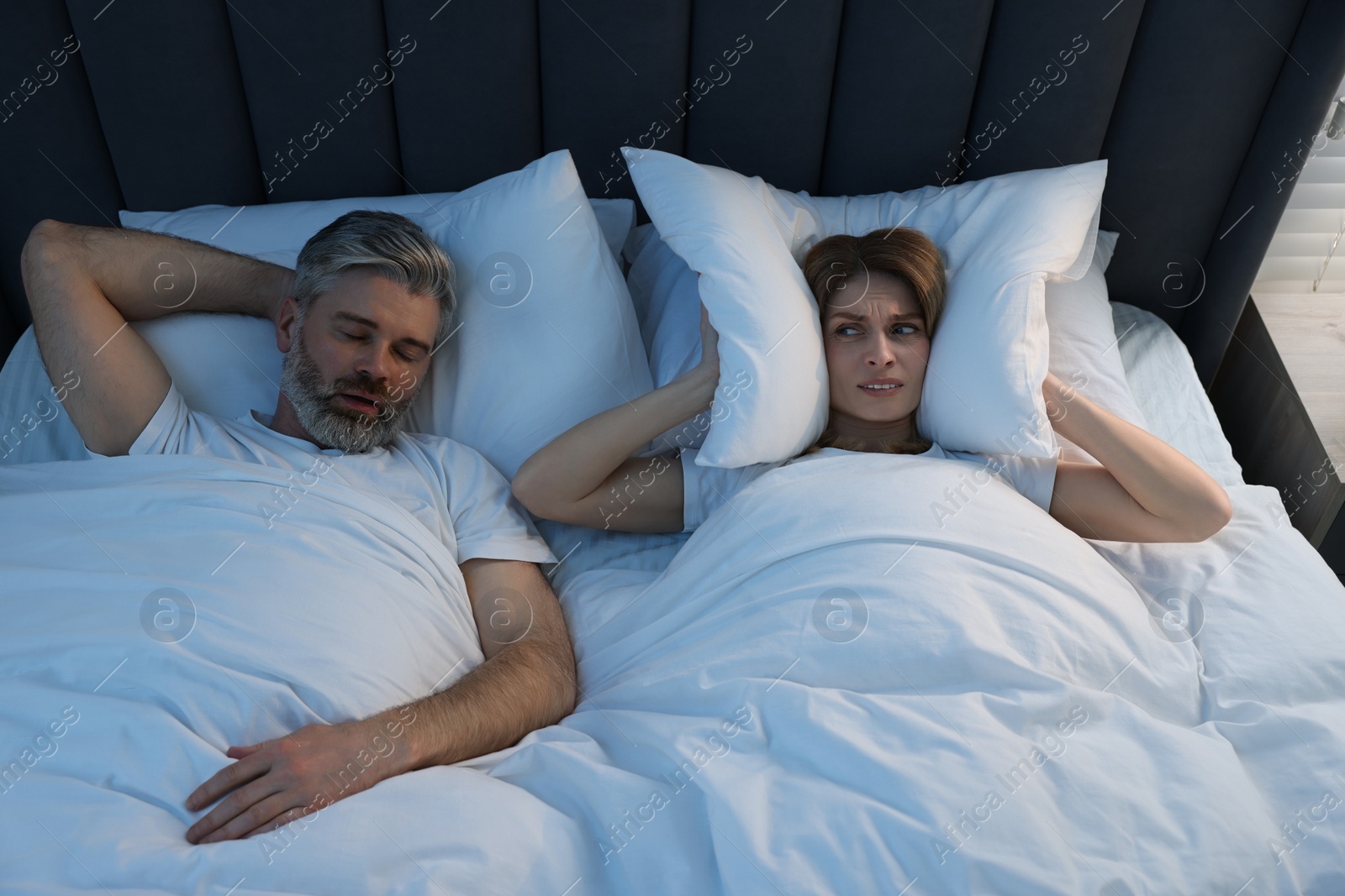 Photo of Irritated woman covering her ears with pillows in bed at home. Problem with snoring husband