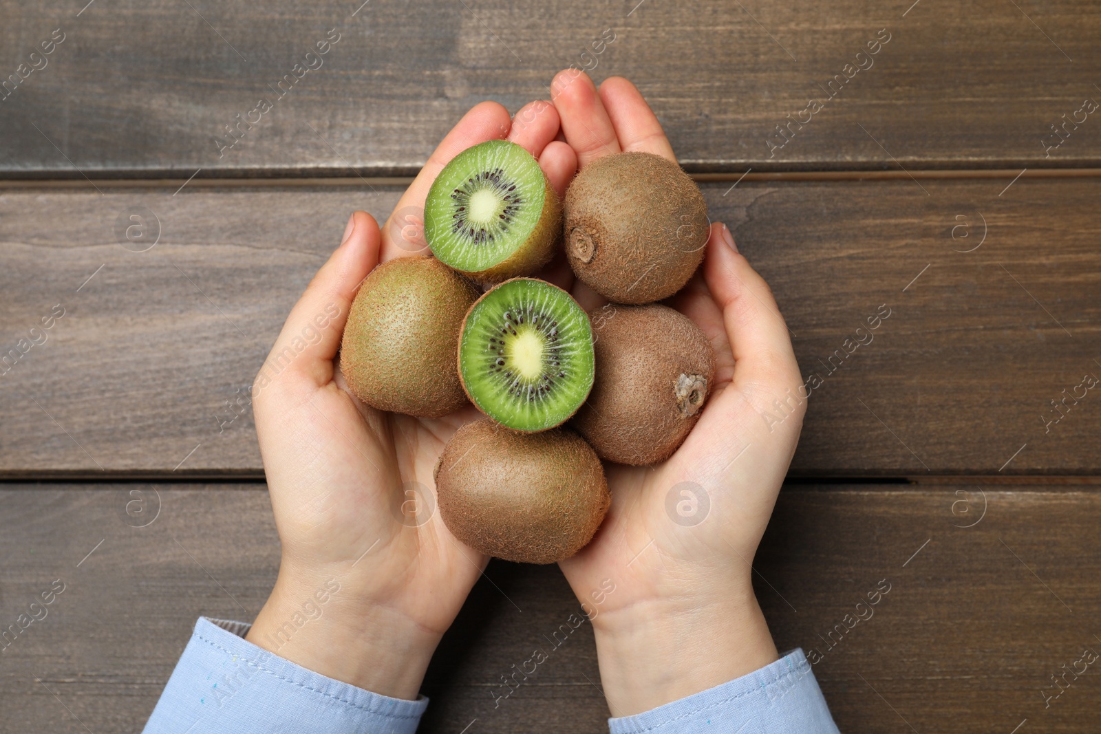 Photo of Woman holding pile of fresh ripe kiwis at wooden table, top view