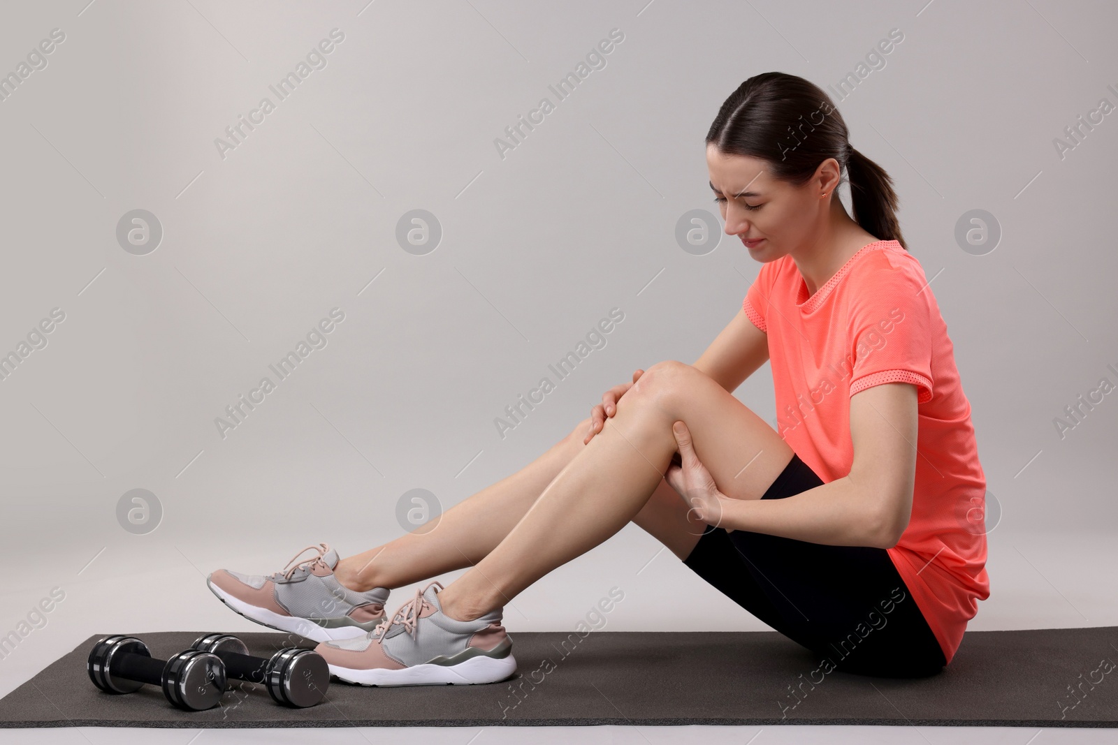 Photo of Young woman suffering from leg pain on exercise mat against grey background