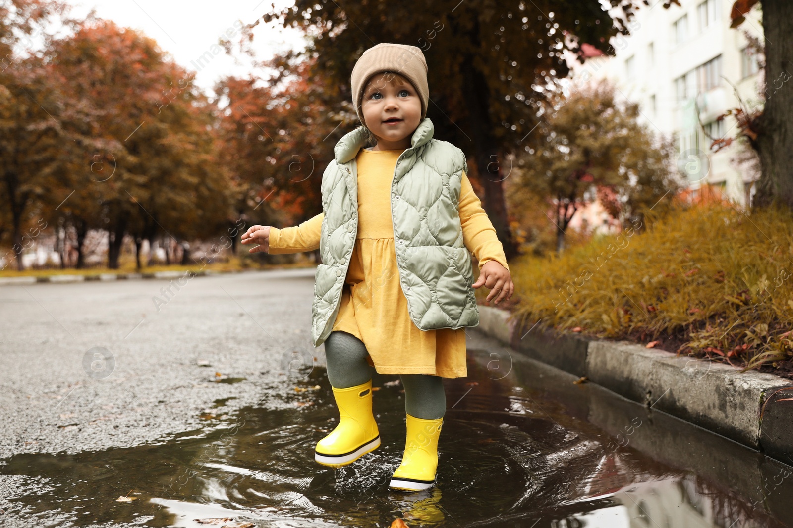 Photo of Cute little girl splashing water with her boots in puddle outdoors