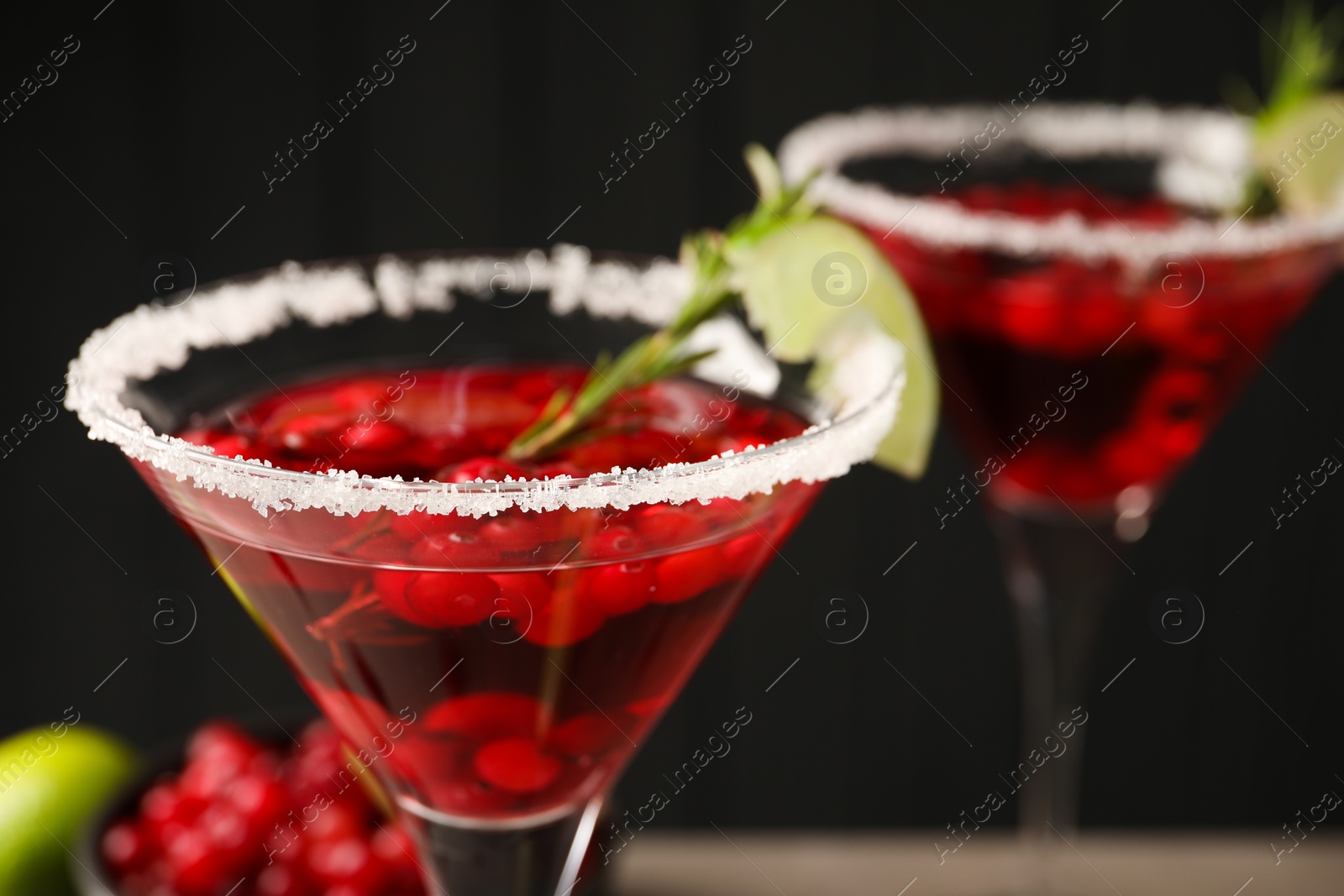 Photo of Tasty cranberry cocktail with rosemary and lime in glasses against black background, closeup