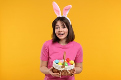 Easter celebration. Happy woman with bunny ears and wicker basket full of painted eggs on orange background