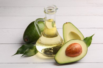 Photo of Glass jug of cooking oil and fresh avocados on white wooden table