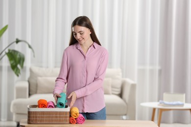 Smiling woman putting rolled shirt into basket at table in room, space for text. Organizing clothes