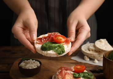 Photo of Woman holding puffed rice cake with prosciutto, tomato and basil at wooden table, closeup