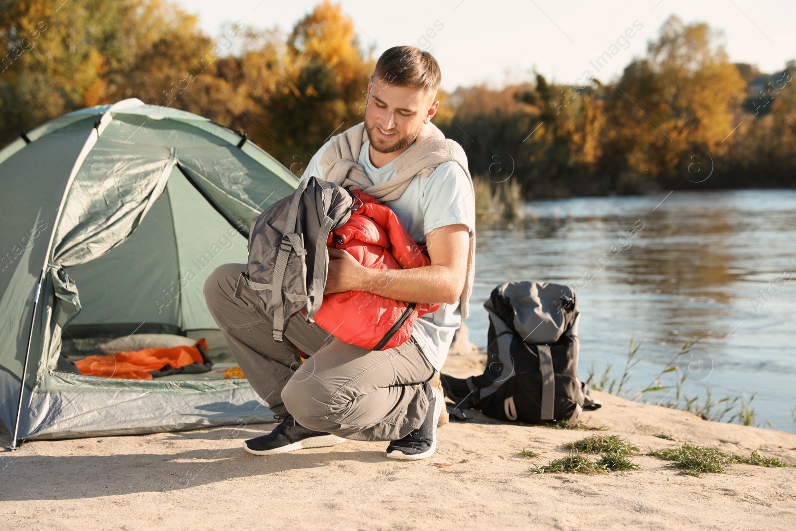 Photo of Young man packing sleeping bag near camping tent outdoors