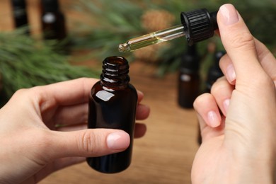 Photo of Woman holding pipette with pine essential oil over bottle at wooden table, closeup