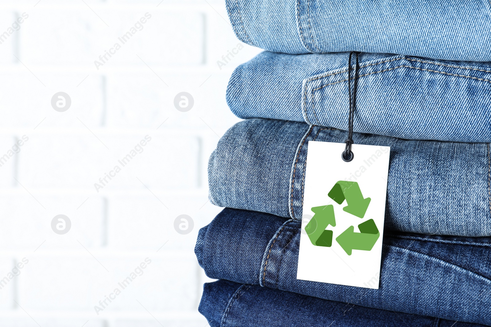 Image of Stack of different jeans with recycling label near white brick wall, closeup. Space for text