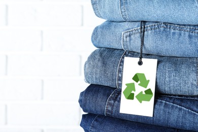 Image of Stack of different jeans with recycling label near white brick wall, closeup. Space for text