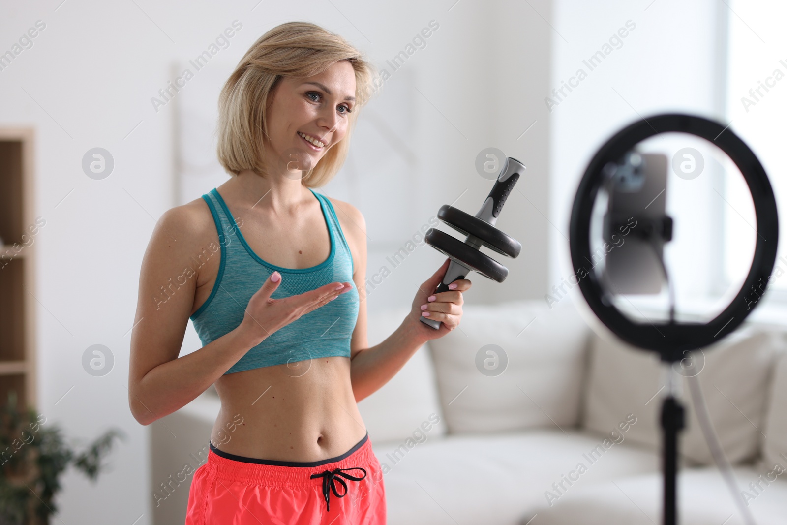 Photo of Smiling sports blogger holding ab roller while streaming online fitness lesson with smartphone at home