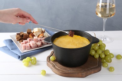 Woman dipping piece of bread into fondue pot with tasty melted cheese at white wooden table against gray background, closeup