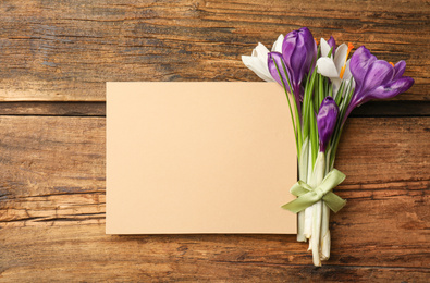 Photo of Beautiful spring crocus flowers and blank card on wooden table, flat lay. Space for text