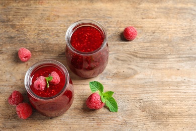 Photo of Glass jars of sweet jam with ripe raspberries and mint on wooden table, above view. Space for text