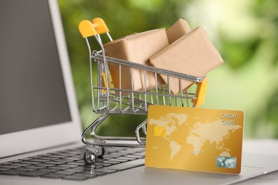 Photo of Online payment concept. Small shopping cart with bank card, boxes on laptop, closeup