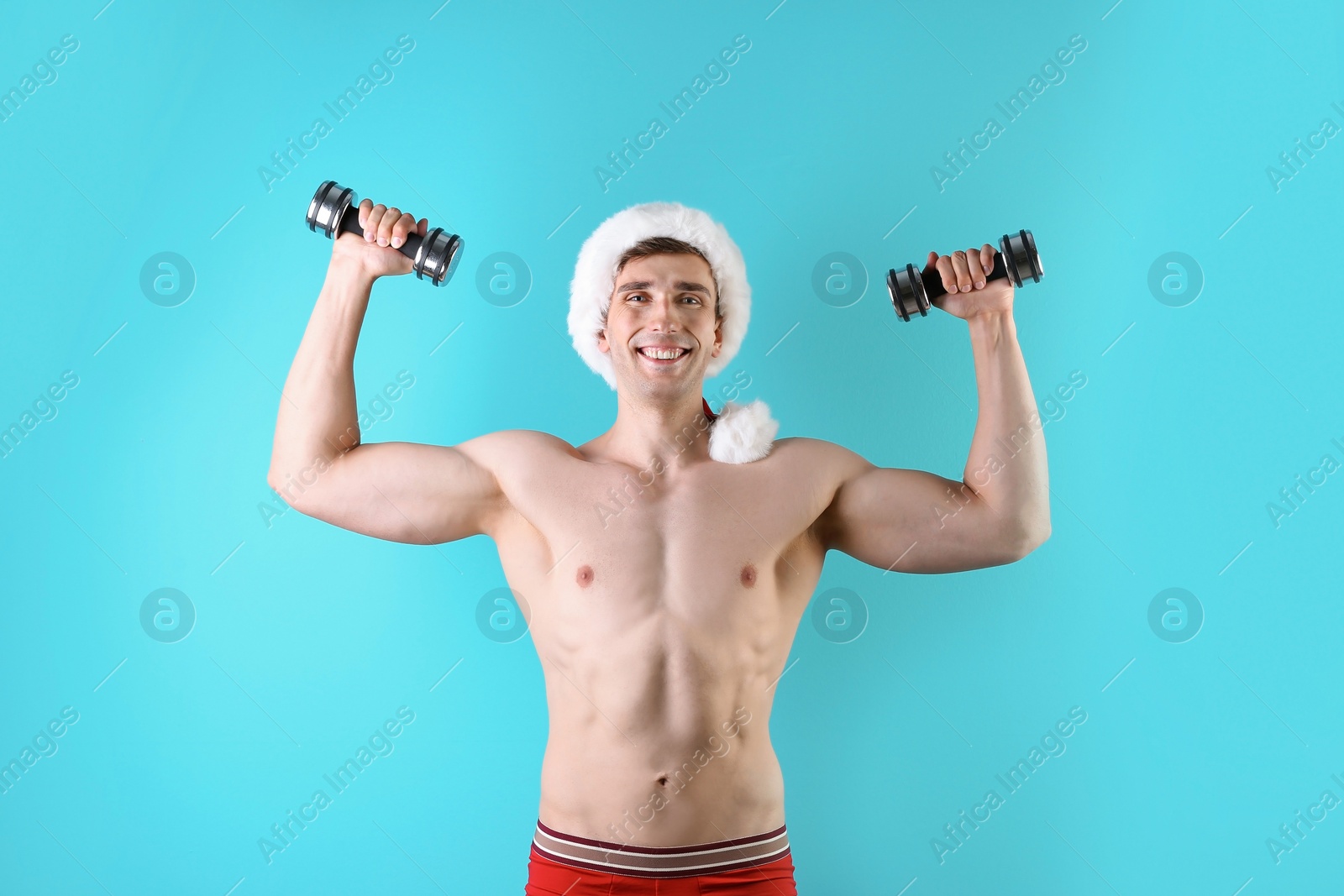 Photo of Young muscular man in Santa hat with dumbbells on color background