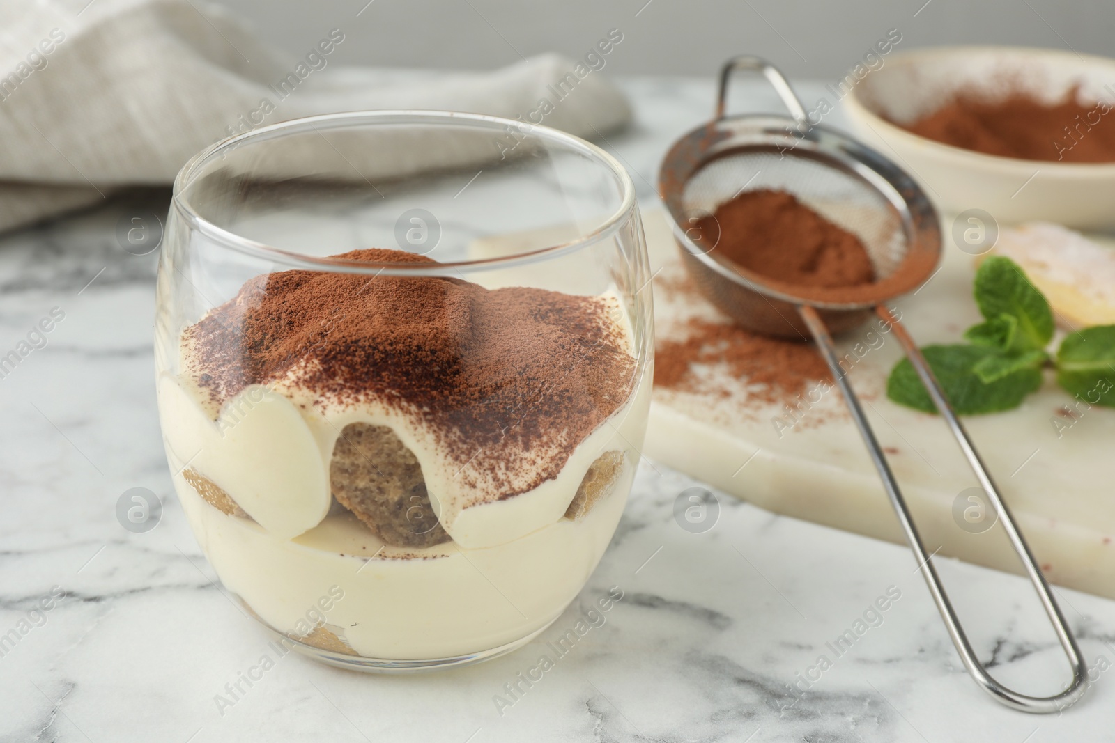 Photo of Delicious tiramisu in glasses and sieve with cocoa powder on white marble table, closeup