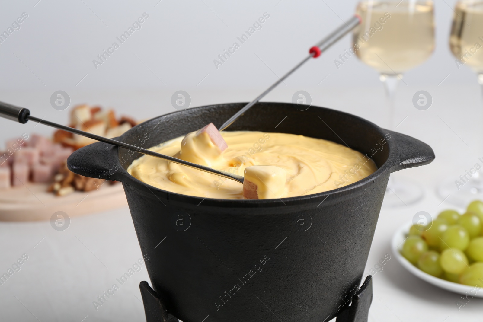 Photo of Dipping ham into fondue pot with tasty melted cheese at white table, closeup