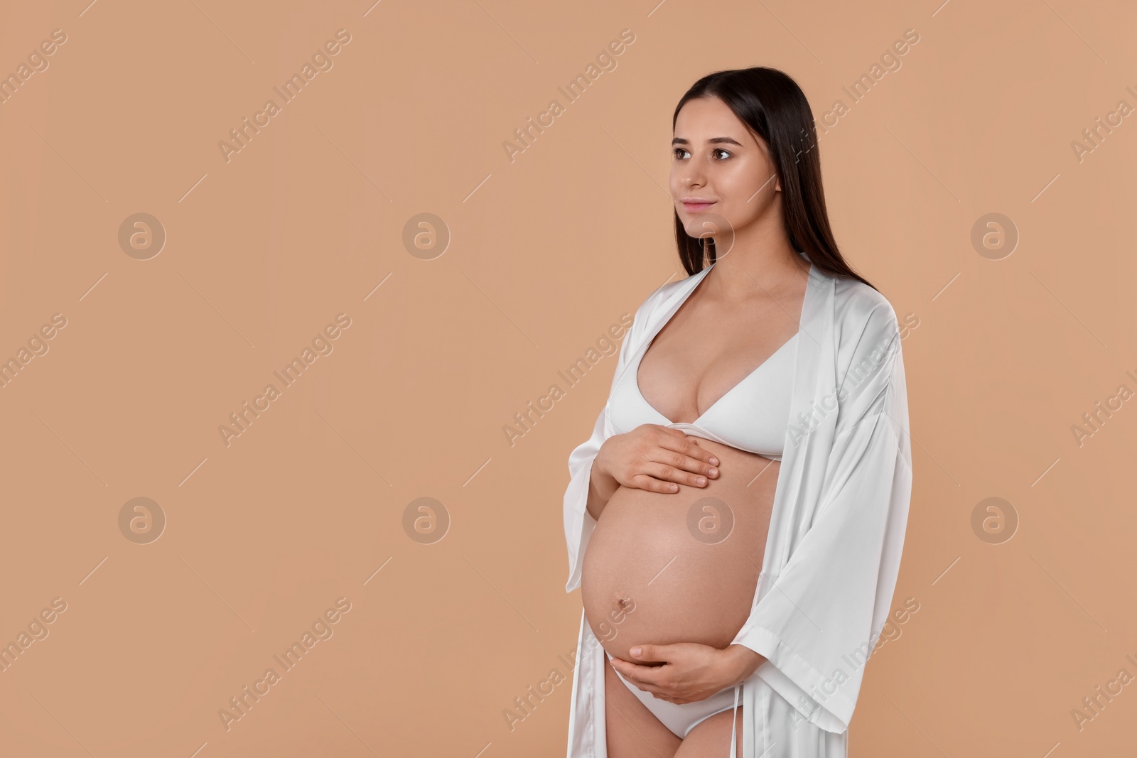 Photo of Beautiful pregnant woman in stylish comfortable underwear and robe on beige background, space for text