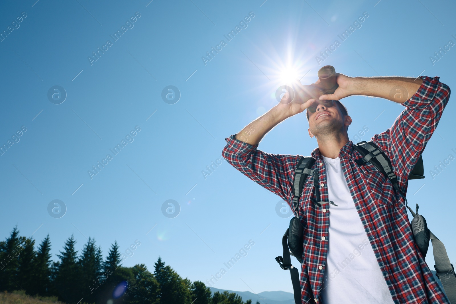 Photo of Tourist with hiking equipment looking through binoculars outdoors on sunny day, low angle view