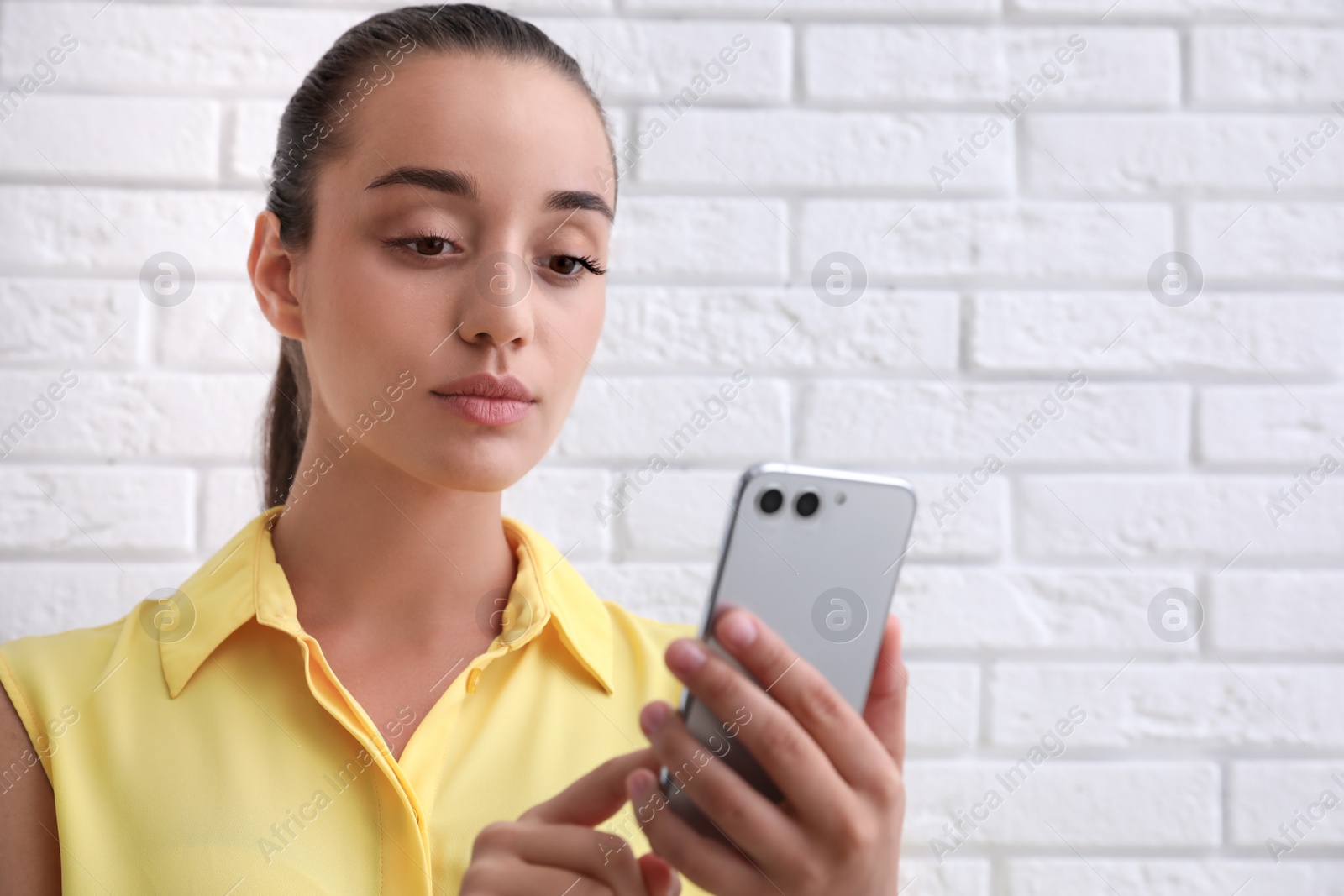 Photo of Young woman unlocking smartphone with facial scanner near white brick wall. Biometric verification