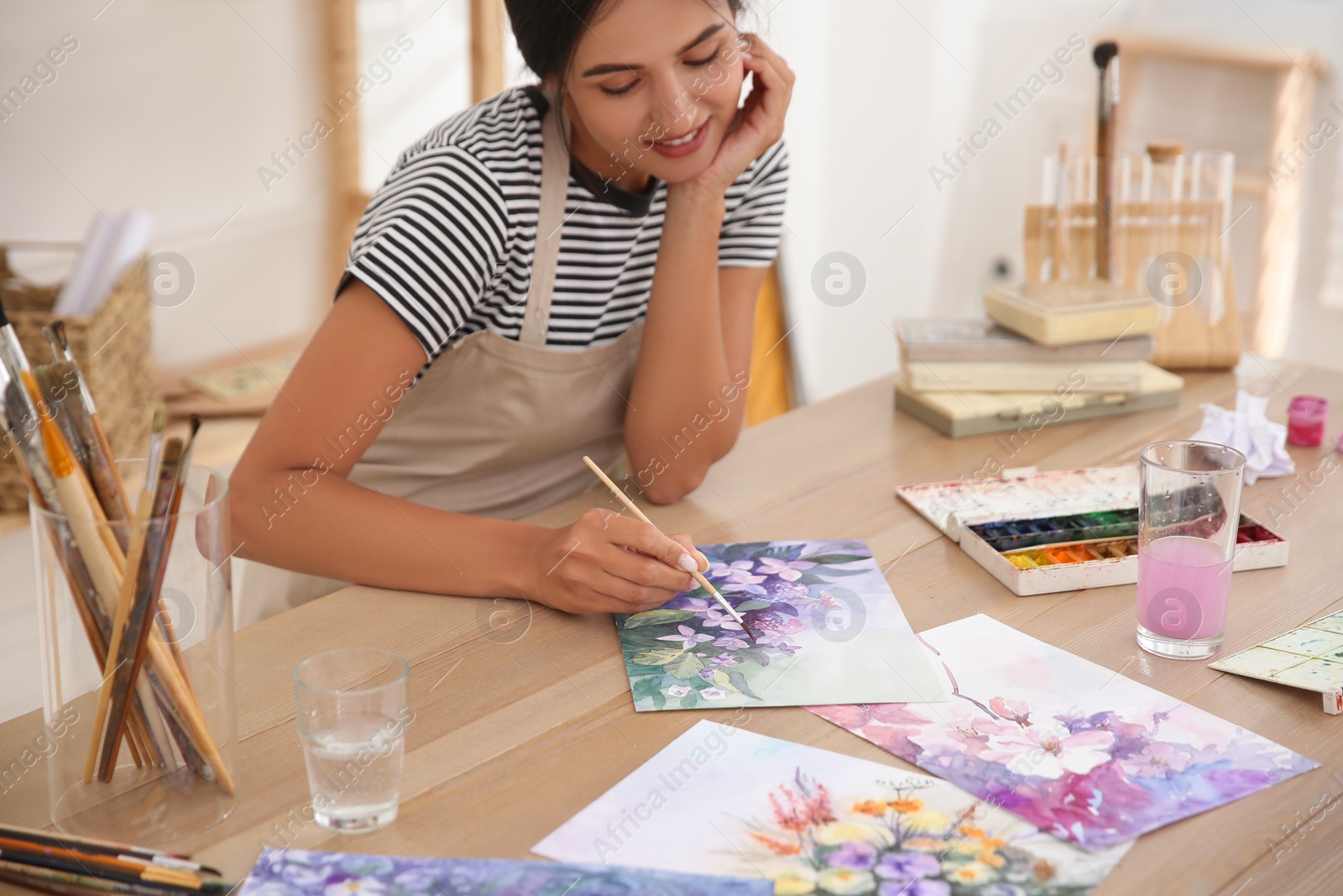 Photo of Young woman drawing flowers with watercolors at table indoors, focus on painting