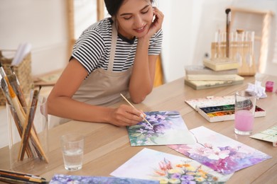 Photo of Young woman drawing flowers with watercolors at table indoors, focus on painting