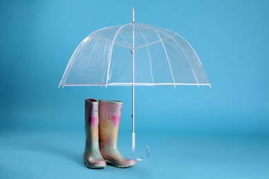 Photo of Open transparent umbrella and rubber boots on light blue background