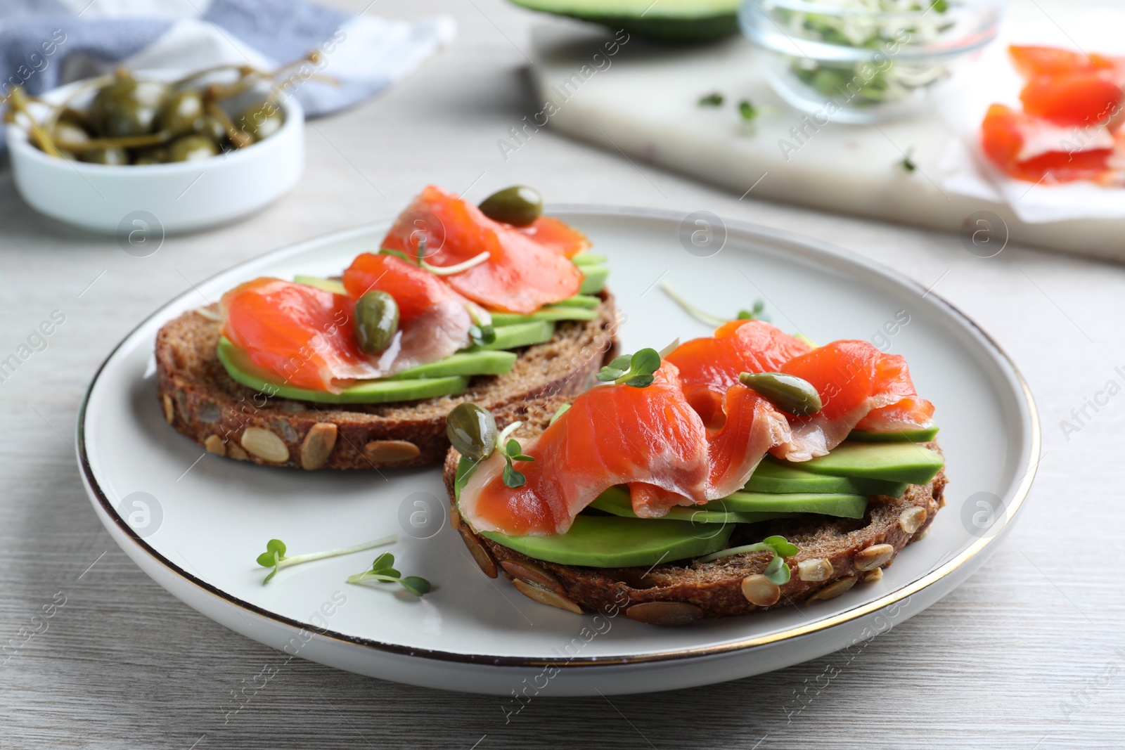 Photo of Delicious sandwiches with salmon, avocado and capers on white wooden table