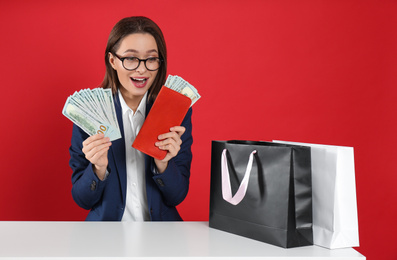 Emotional young woman with money and shopping bags at table on crimson background