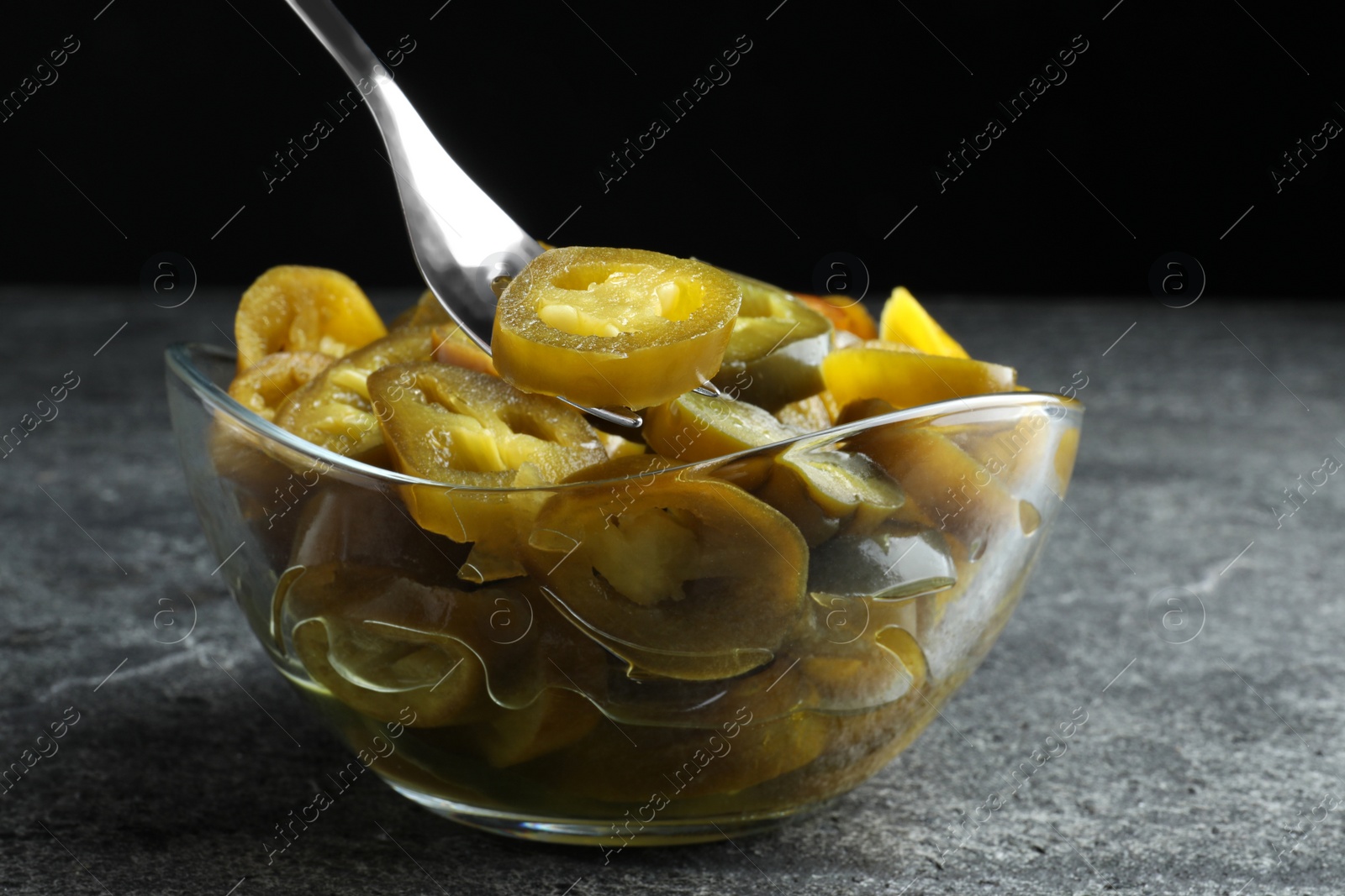 Photo of Taking slice of pickled green jalapeno with fork from bowl on grey table