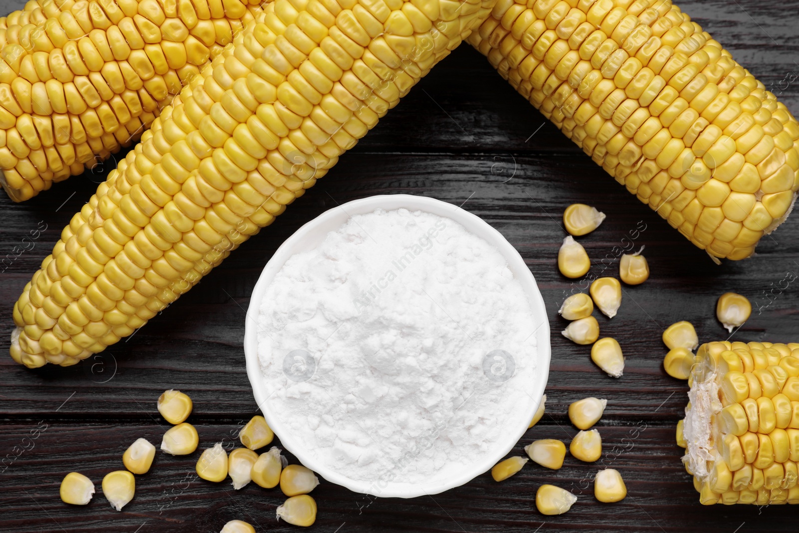 Photo of Bowl with corn starch, ripe cobs and kernels on dark wooden table, flat lay