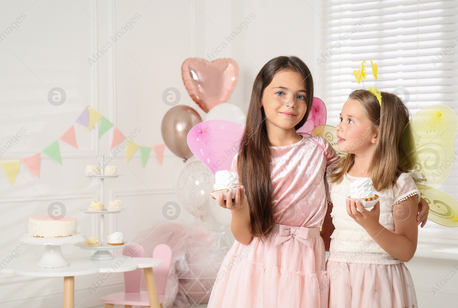 Photo of Cute little girls wearing fairy costumes with delicious cupcakes in decorated room. Space for text