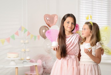 Photo of Cute little girls wearing fairy costumes with delicious cupcakes in decorated room. Space for text